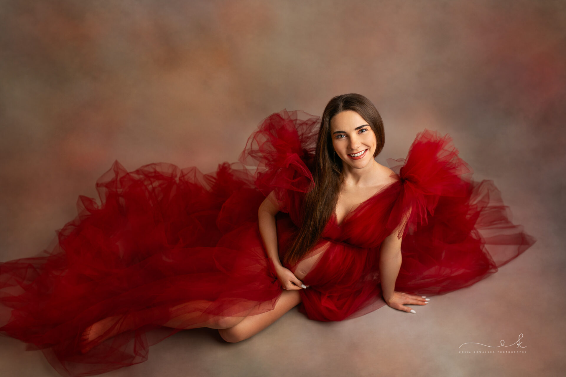 artystyczna fotografia ciążowa, sesja w tiulach, mama siedzi na ziemi w bordowej tiulowej sukni - sesja studyjna Kraków