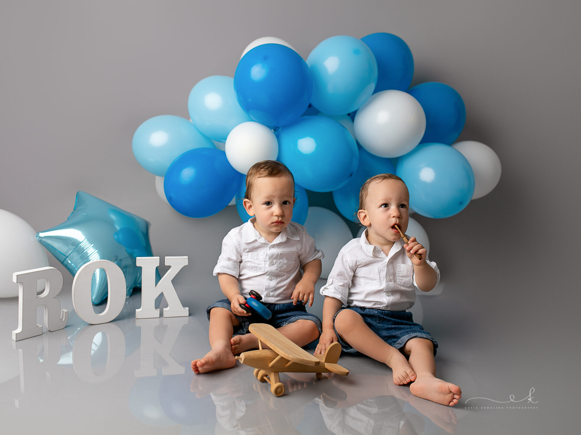 Pierwszy rok bliźniąt, sesja na roczek, niebieskie i białe balony w tle, napis rok, chłopcy bawią się samolotem i samochodzikiem